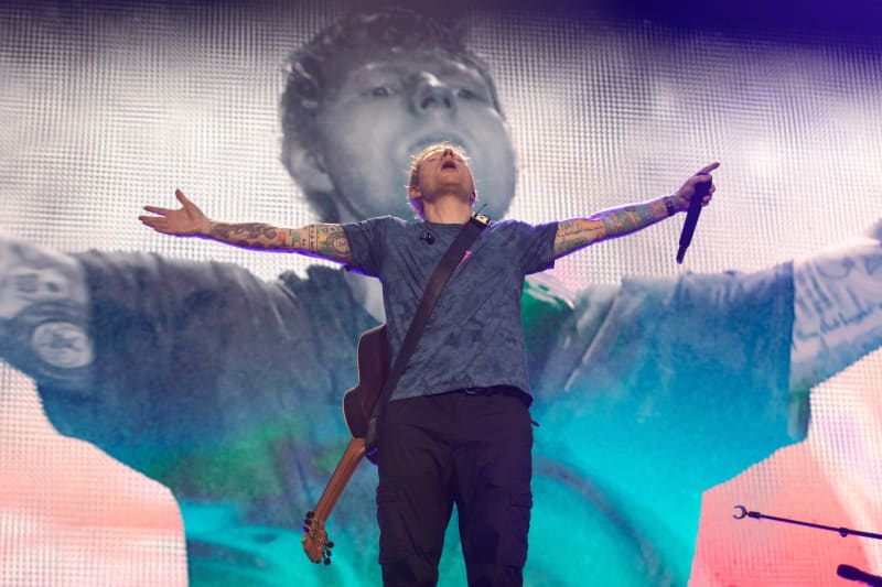 British pop star Ed Sheeran on stage with his arms wide open, mic in left hand and guitar slung on shoulder.