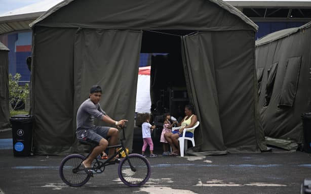 A resident rides a bicycle past a temporary shelter housing people displaced by the twin earthquakes at the Jose Maria Vargas sports complex in Maiquetia, La Guaira state, Venezuela, on July 13, 2026. The death toll from the twin earthquakes that struck Venezuela nearly three weeks ago has surpassed 4,500, while the number of injured remains at about 17,000, according to the latest government report released on July 13. (Photo by Federico PARRA / AFP)