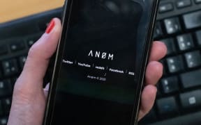 An illustration picture shows the AN0M logo displayed on the screen of a smartphone.