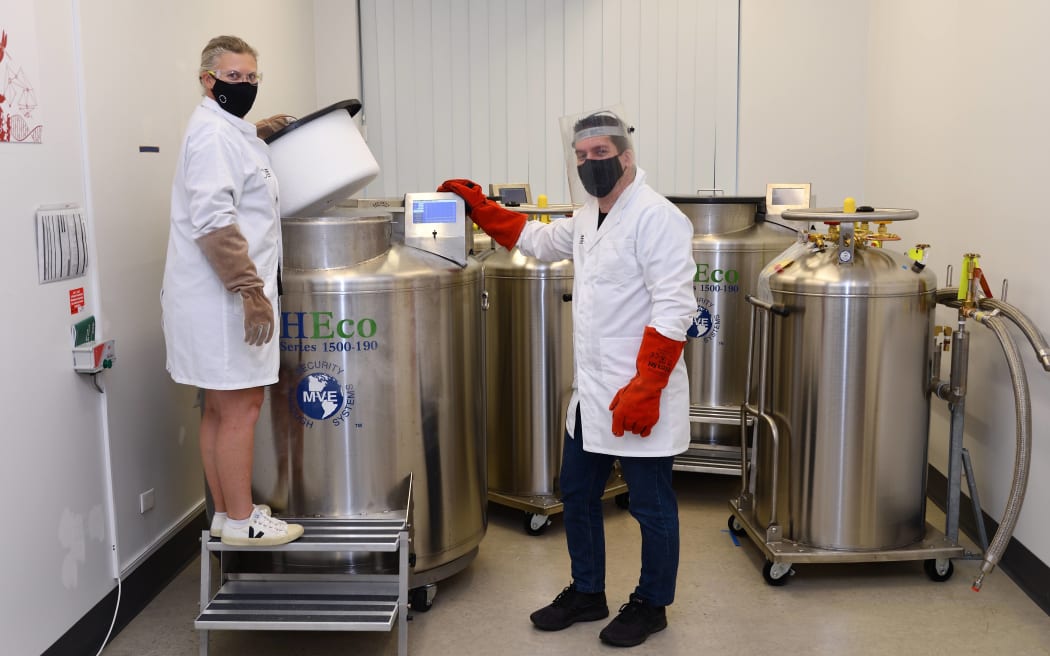 Megan stands on a step in front of a large liquid nitrogen tank - she is wearing a lab coat, thermal gloves, goggles and a mask. She has lifted up the lid of the tank. Bevan stands to the right of the tank also in labcoat, gloves and a face shield, his right hand rests on the tank display. The tank is shiny stainless steel & comes up to Bevan's shoulder height.