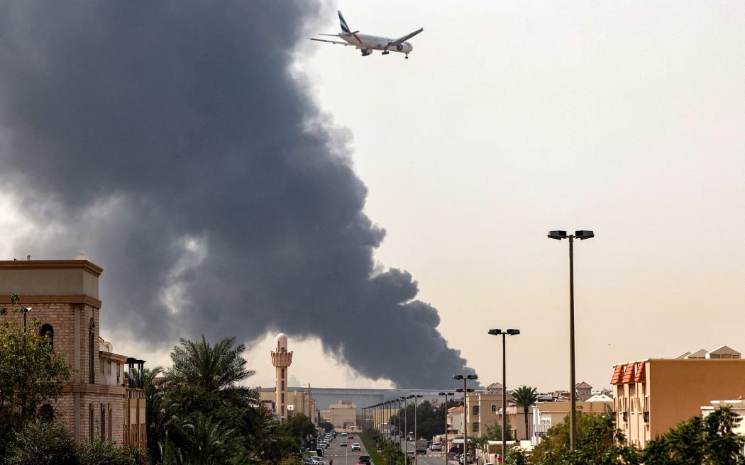 An Emirates aircraft prepares for landing as a smoke plume rises from an ongoing fire near Dubai International Airport in Dubai on March 16, 2026. Flights were gradually resuming at Dubai airport on March 16, previously the world's busiest for international flights, the airport operator said, after a "drone-related incident" sparked a fuel tank fire nearby, as Iran kept up its Gulf attacks.