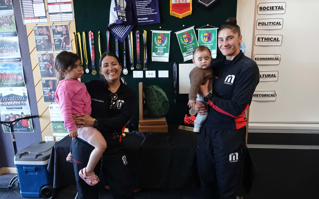 Kristina Sue, left, and Rhiarna Ferris with their daughter, Makereti, 4, in pink, and son, Māhaki, 6 months, in front of the national title trophy and wall of accolades.