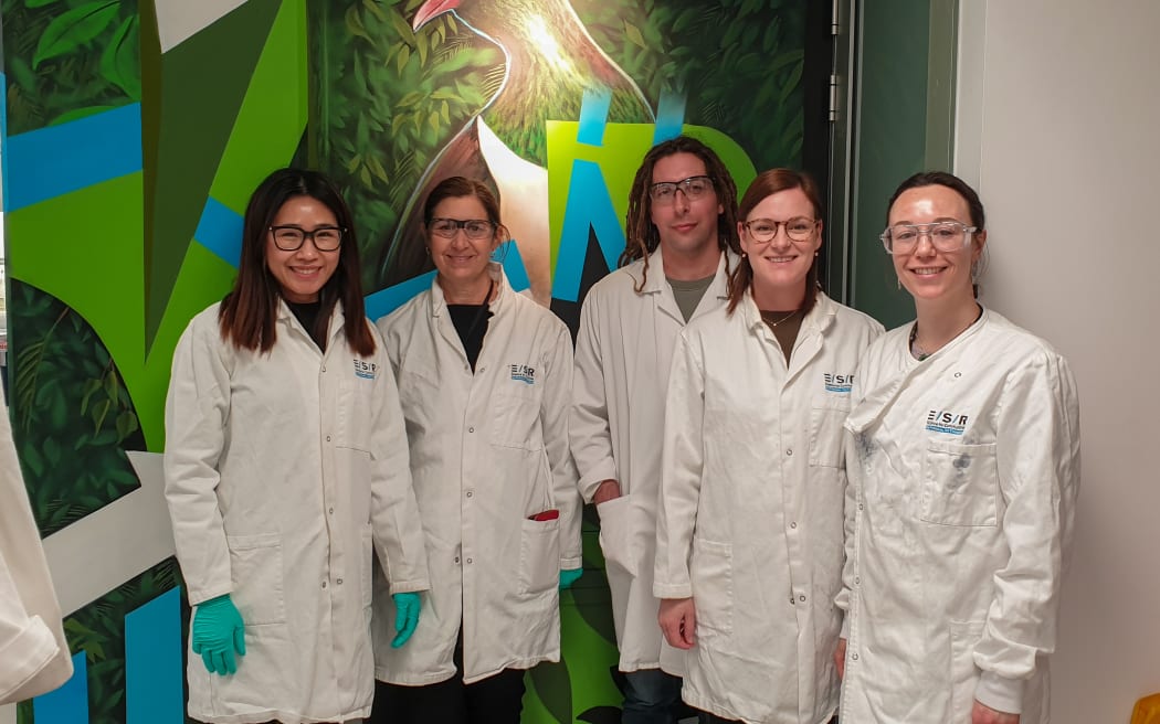 Five people wearing white lab coats and goggles stand in front of a mural of a kererū.