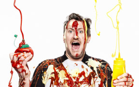 Max halley wears a napkin around his neck. In his left hand he is holding some tomato ketchup, in his left a tube of mustard. He is spraying their contents into the air. Max is covered in streaks of ketchup and mustard. His expression is happy and joyful.