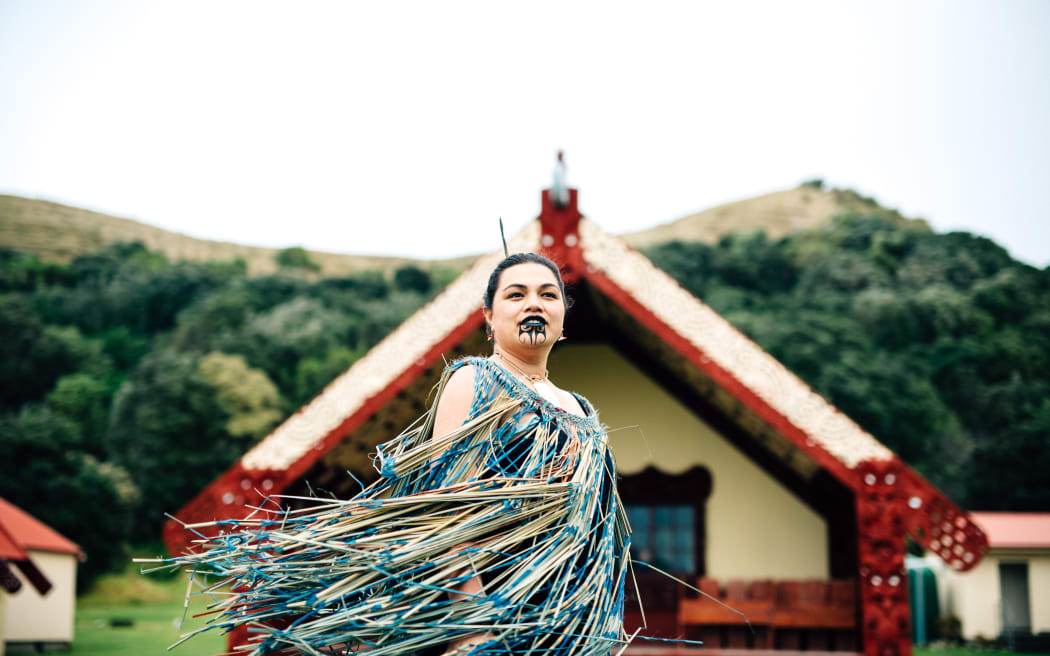 Ruth stands out the front of a marae, her clothes blowing in the wind.