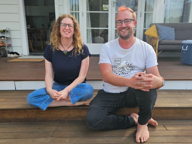 Two friends, a woman and a man are sitting side by side on the outside steps of a deck, smiling at the camera. They are barefoot, casually dressed and look relaxed.