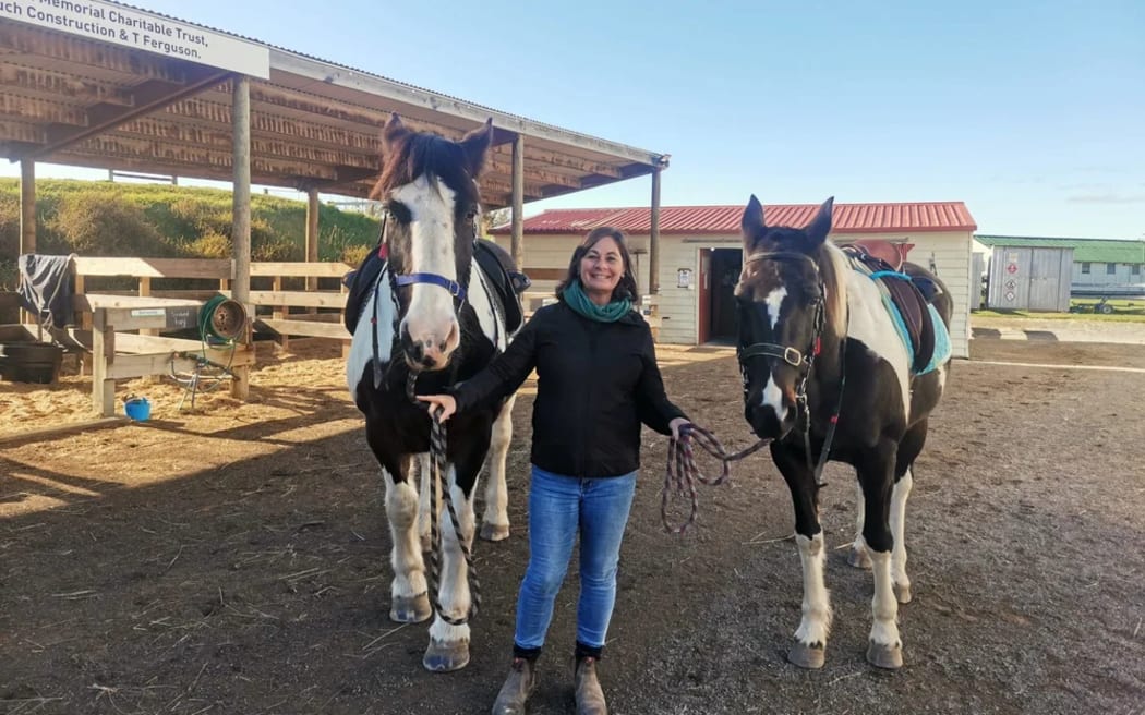 Jo stands by some stables holding two horses by their reins. She smiles.