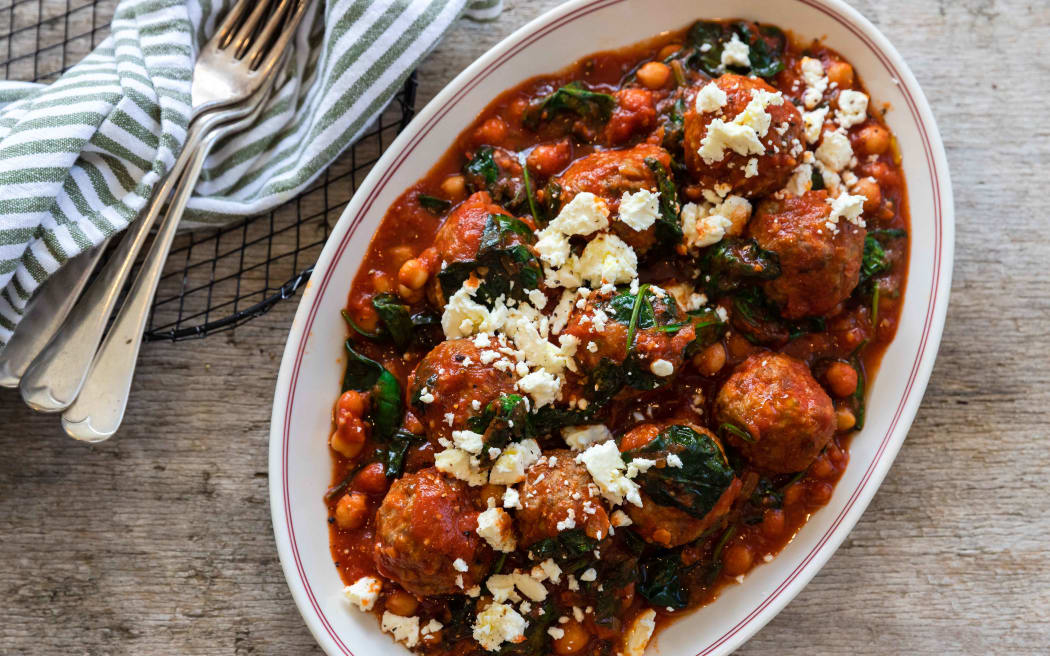 An oval dish on a wooden board containing Spanish meatballs with chilli, chickpea and spinach sauce