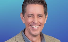 Host Wallace Chapman in a blue shirt and casual jacket.Soft blue background.