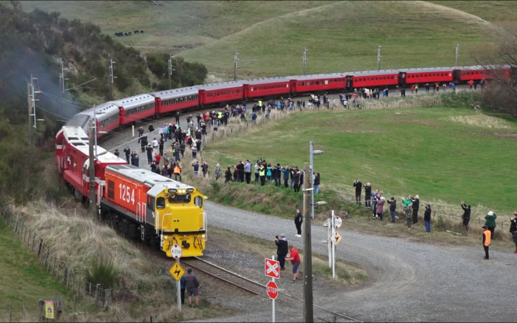 A train glides on the rail tracks around a bend, spectators are outside taking photos and watching the train go past a few meters away from the train track.