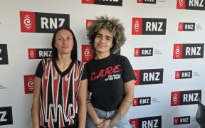 Two women stand against a wall that reads RNZ.