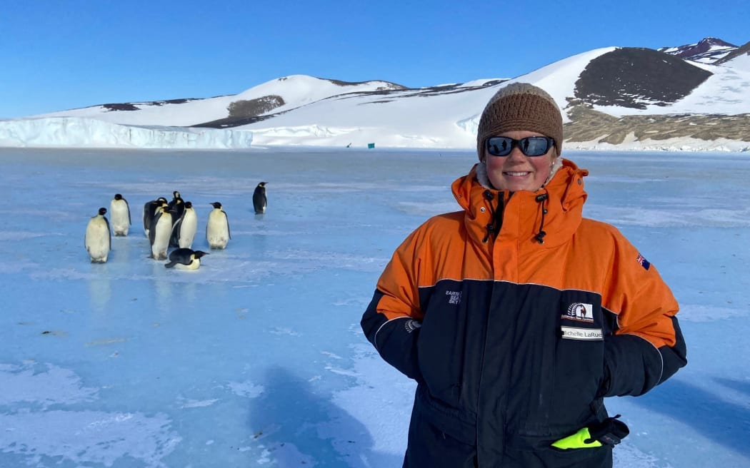 A mid-shot of a woman wearing sunnies, a beanie and black-and-orange warm jacket standing on the ice in front of a small huddle of about 10 emperor penguins. Behind them is a snow-blanketed rocky hill.