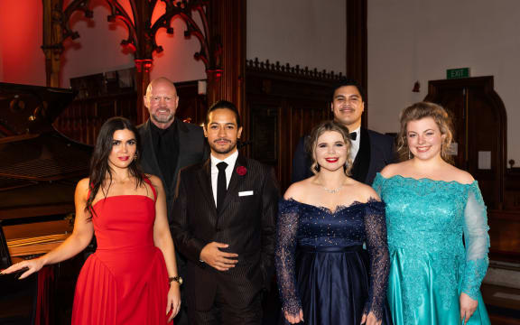 six opera singers on stage in formal ware, smiling at the camera.