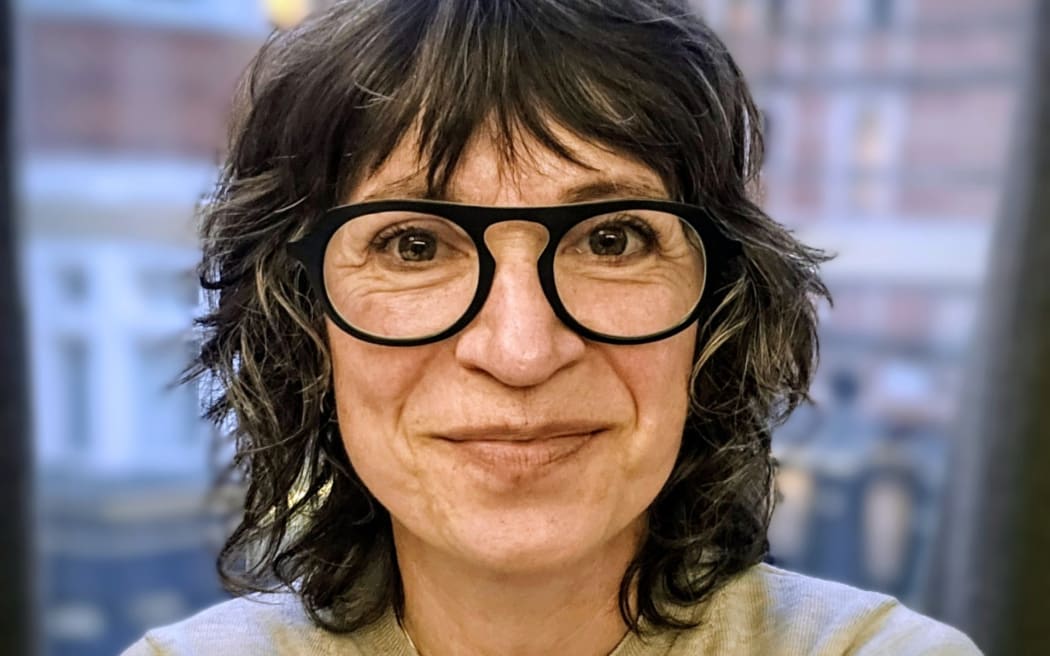 This is a headshot of Cass. She is in front of a window. It's impossible to tell where the photo is taken but there are some brick buildings behind her that look a bit like London. It could also be NZ though.