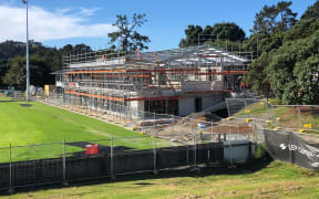A large building is being constructed next to a green sports field. The structure is covered in scaffolding.
