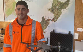 Jordan is wearing an orange high vis top in front of a map with New Zealand. He is holding a quad drone in his left hand, in front of the open padded box that it is stored in.