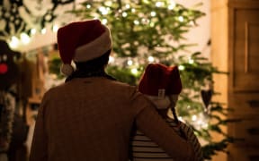 An adult and a child wearing Christmas hats sitting in front of a Christmas tree with their backs to the camera.