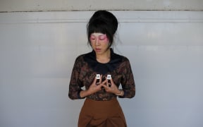 A woman in a brown shirt with a black collar stands in front of a white wall. She holds up two white cubes inscribed with Chinese characters.