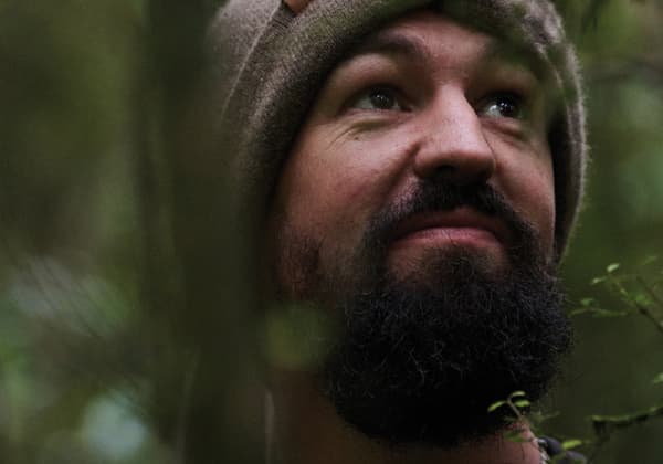 Sam Gibson, a bearded man wearing a beanie, looks to the sky with forest behind him.