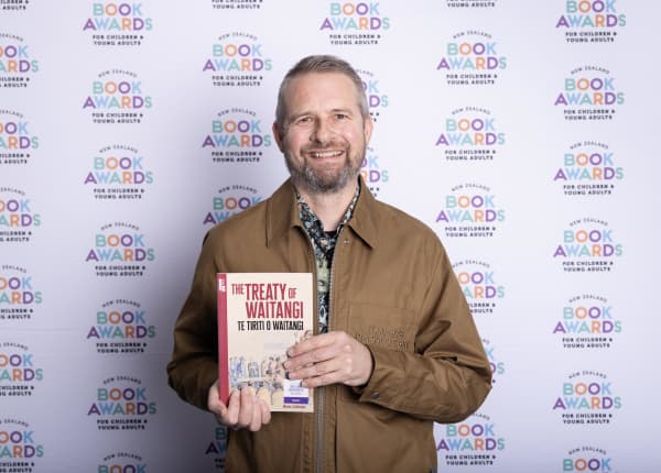 Author Ross Calman, wearing a brown zip up jacket, holds his book, The Treaty of Waitangi.