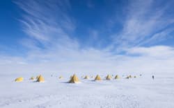 A shot of the Ross Ice Shelf which is flat and white to the horizon, with a blue, feathery-cloud sky. There are a number of yellow tents on the ice, most of them triangle shaped, with a few that are flatter and rounder. One small figure is walking towards the tent, leaning against the wind, which is obviously strong by the strung out flag in the foreground.