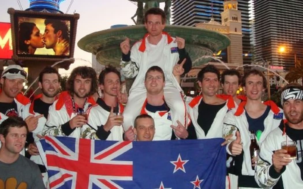 Geoff 'The Nimble Thimble' Christopher sits on his team mates shoulders in front of a fountain at the 2009 Monopoly World Championship in Las Vegas. They are holding a big New Zealand flag in front of them and wearing a matching team kit which also features the flag.