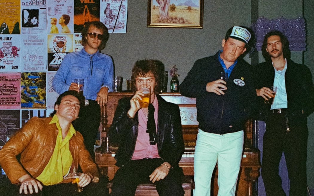 This is a 70s style image of the band in a country pub or possibly hall. The pub has brown wooden floorboards and posters on the wall. They are stood in front of an old piano. A slightly crooked painting of a tree hangs on the wall behind the piano. If I had to guess i'd say it was an Australian pub. Zooming in confirms this as the postersi in the background make it clear it's the Fitzroy Pinnacle in Melbourne.