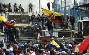 A demonstrator waves an Ecuadorean national flag while police officers stand guard in the surroundings of the National Assembly in Quito on June 25, 2022, when a parliamentary session called by opposition lawmakers for a no confidence vote against President Guillermo Lasso will be held in the framework of indigenous-led protests against the government. - An estimated 14,000 protesters are taking part in a nationwide show of discontent against rising hardship in an economy dealt a serious blow by the coronavirus pandemic. Protesters are demanding a cut in already subsidized fuel prices, which have risen sharply in recent months, as well as jobs, food price controls, and more public spending on healthcare and education. (Photo by Rodrigo BUENDIA / AFP)