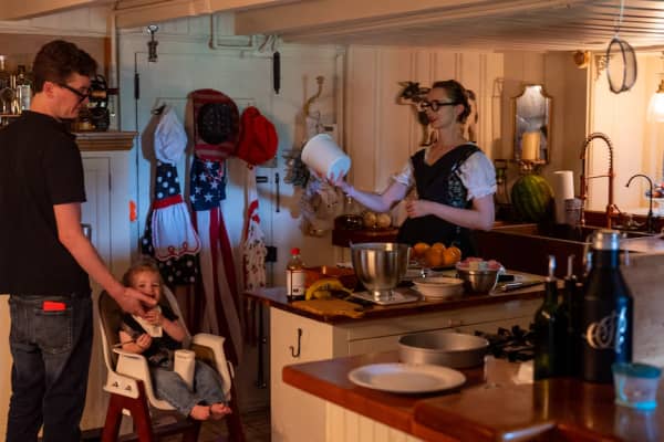 Malcolm Collins stands near his toddler sitting in a baby chair in their kitchen. Simone stands nearby holding a container.