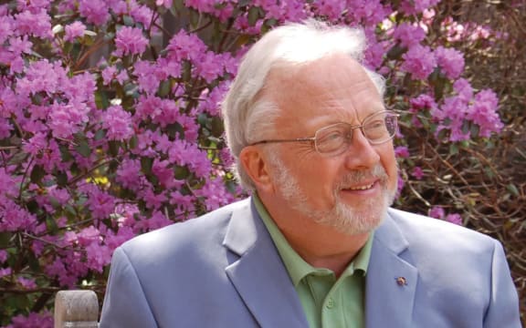 American composer William Bolcom, seated in front of a lilac azalea bush