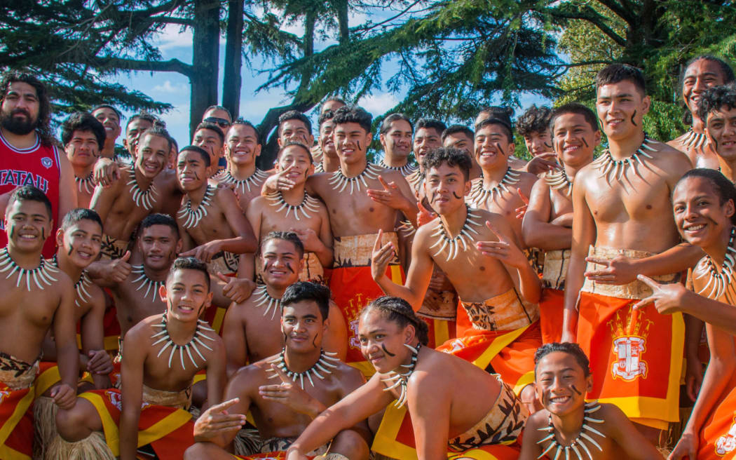 A large group of smiling young performers in traditional Polynesian attire pose together outdoors. They wear orange and yellow garments, necklaces made of white spikes, and have black markings on their faces. Above them, on a yellow banner, bold black text reads “SEVEN WEEKS,” with the subtitle “JOURNEY TO POLYFEST” in italics.