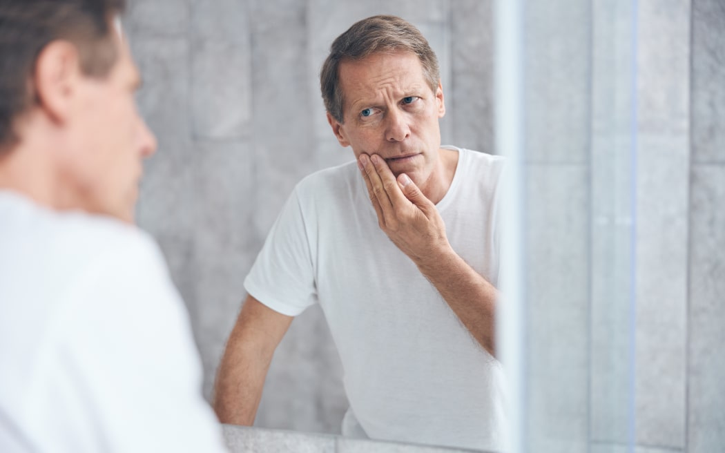 Waist-up portrait of a low-spirited male with his hand on the cheek looking at himself