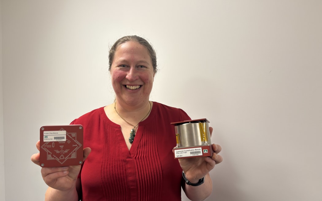 A person wearing a red shirt is holding two objects. The object in the left hand is a red square device with white markings and a barcode. The object in the right hand is a cylindrical device with gold and silver components.
