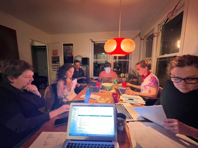A group of adults gathered in a lounge on a dining table, each with paper work and their laptops out.