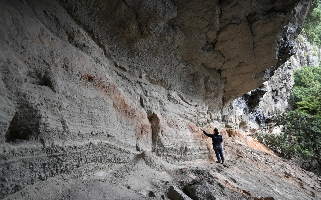 Jacqueline stands in the midground of the picture in a big overhang of grey volcanic rock, with an orange layer running through it.