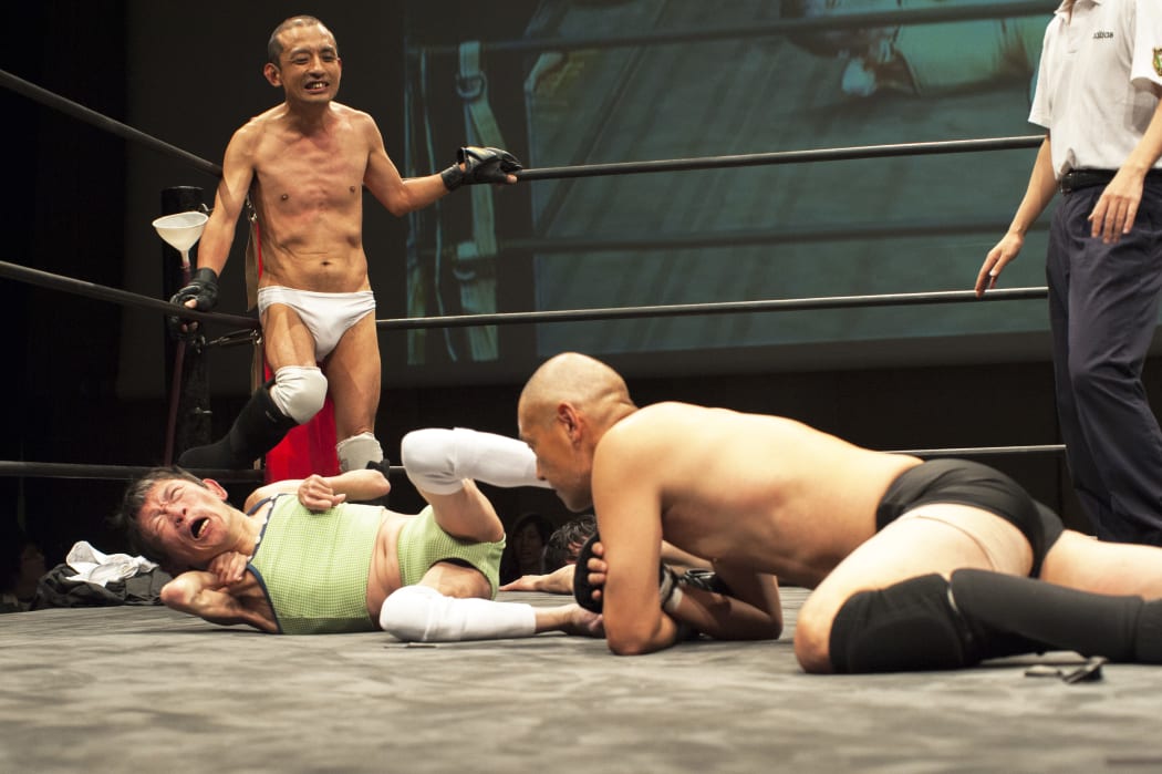 Shintaro Yano ( "Sambo" Shintaro) watches on as tag-team partner L'Amant delivers a kick to able-bodied "Antithesis" Kitajima's face.