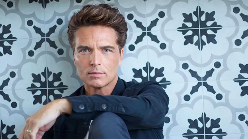 Singer Richard Marx wearing a blue shirt and with fabulous bouffant hair sits in fron of a blue patterned tiled wall.