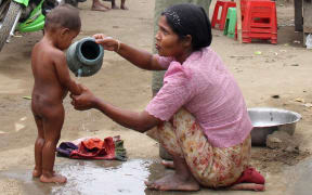 A mother tips water over a toddler who stands naked on a dirt path