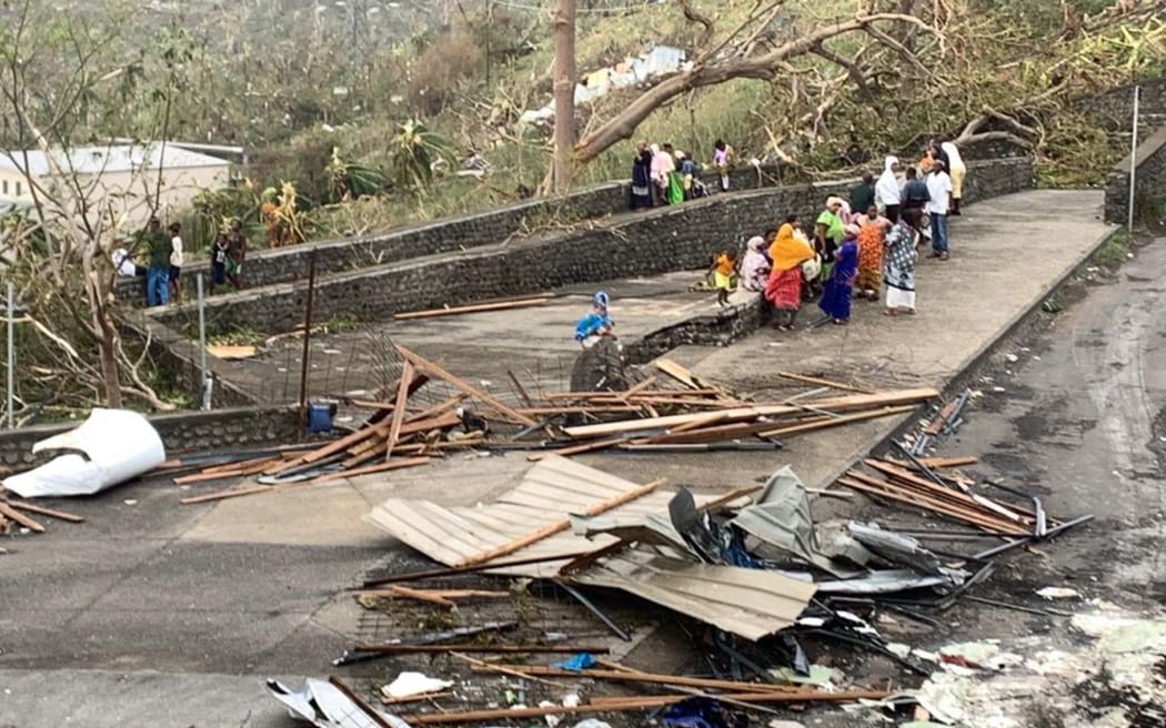 A photo taken on December 15, 2024 shows residents sitting by a road among piles of debris of metal sheets and wood after the cyclone Chido hit France's Indian Ocean territory of Mayotte. - At least 14 people were killed in Mayotte when a fierce cyclone battered the French Indian Ocean territory, authorities said on December 15, 2024, with officials warning it will take days to know the full toll. (Photo by KWEZI / AFP)