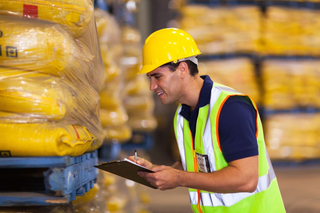 A warehouse worker wearing a high-vis vest holds a clipboard (file).