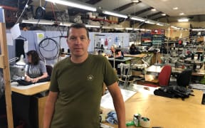 Mat Junge stands in front of a workbench in the Albion Clothing factory. There are other workers at workbenches behind him using sewing machines and other tools. The factory floor is busy with equipment and goods.