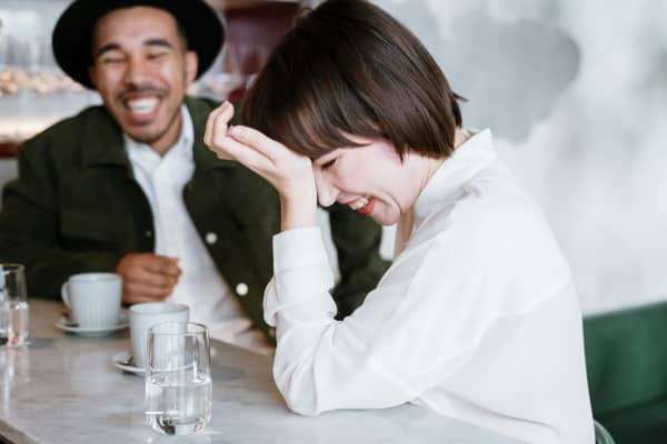 A dark-haired woman in a white shirt laughs with a smiling bearded man in a black hat.