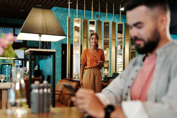 An attractive woman in an orange shirt carrying a phone approaches a man in a bar.