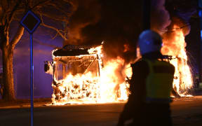 An anti-riot police officer standing next to a city bus burning during unrest in Malmo, Sweden.