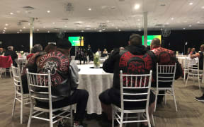 Patched Mongrel Mob members sitting around a table in a conference venue, listening to someone talking on a stage. Their backs are to the camera.