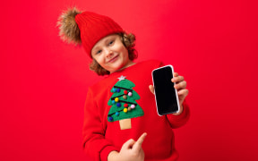 A young boy with a smartphone in his hands against the background of a red wall, in a knitted hat and a sweater with a Christmas tree.