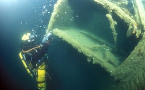 A diver in technical gear shining a light on a big metal structure deep underwater. The metal, part of the RMS Niagara, is covered in a thick layer of rust and plant growth.