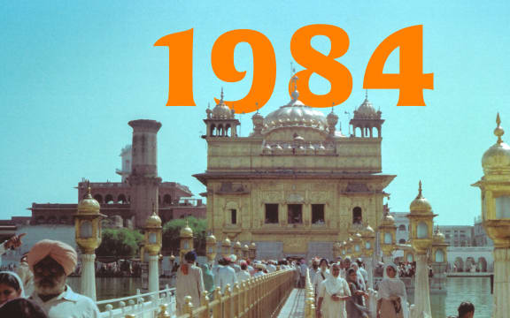 In India, a group of people walk down a path in front of a historic building. Above the building in orange reads: "1984"