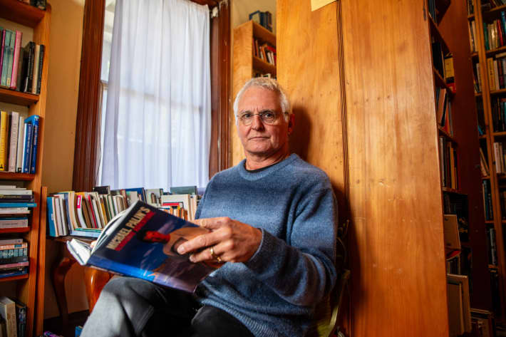 Hard to Find Books shop owner Warwick Jordan sits on a small chair in his shop, with a book open in his hands.