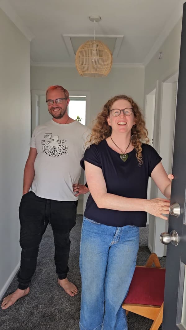Two friends, a man and a woman open their front door, smiling. Behind them you can see the hallway of their new home they have recently purchased as co-owners.